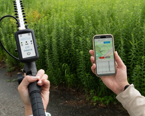 Trained agency staff member holds a tracking antenna and a smartphone displaying a tracking app, surrounded by tall vegetation.