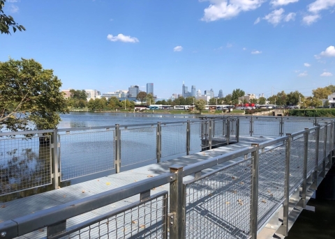 A view of a new pier with metal railings and synthetic decking jutting into a river with a city skyline in the distance