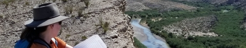 A person wearing a hat stands near a cliff and looks towards look towards a river valley.