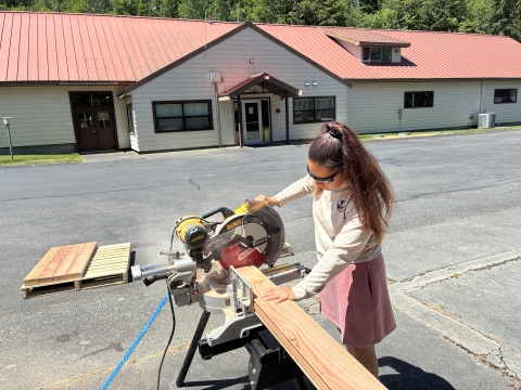 Quilcene National Fish Hatchery intern, Elena Prest, sawing a wooden board with the hatchery facility in the background. 