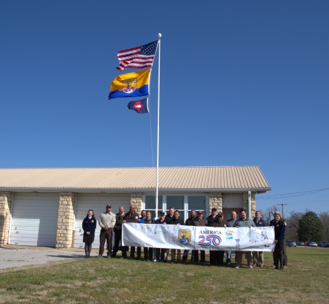 People hold FWS A250 banner at base of flagpole with US flag, FWS flag, fish pennant flying