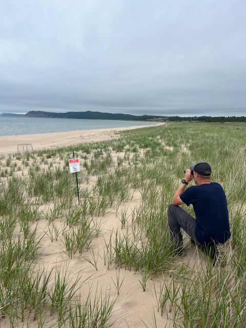 A man kneeing on a beach surrounded by grass looking through binoculars out onto the water.
