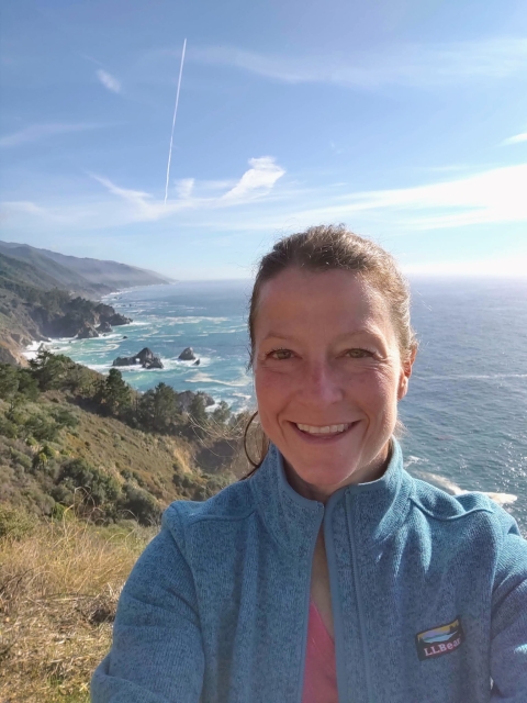 A brown-haired women standing on a cliff with the ocean in the background.