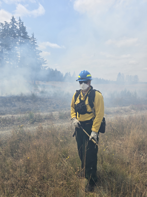 Addison Emerson standing in a field wearing fire safety gear. Black and smoldering field in the immediate background followed by trees and skyline.