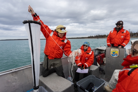 USFWS biologists wearing float coats are on a boat with one explaining and holding a plankton tow net.