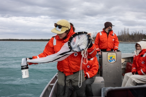 Biologists on a boat wearing float coats. One is holding a plankton tow net on the side to sample for invasive mussels.