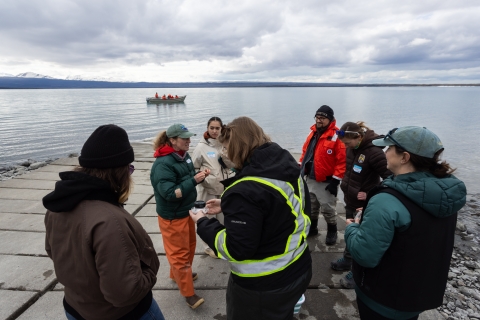 A group of biologist gathered in a circle talking on a lake shore. Calm water and a boat are visible in the background.
