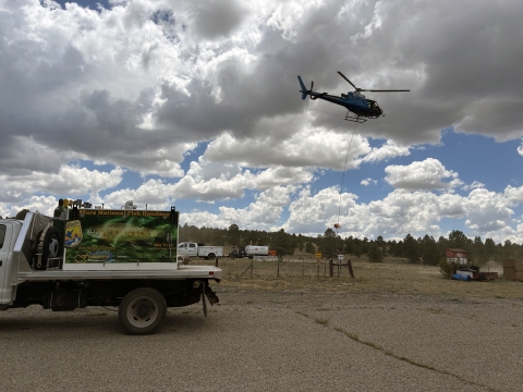 A helicopter hoovers with rope attached to a vessel and above a parking lot on a cloudy day