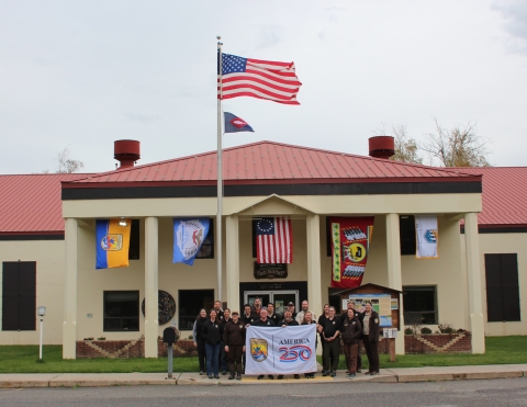 A group of people in brown holding a white “America 250” flag stand in front of a tan building with a red roof. Flags from USFWS, the Confederated Tribes and Bands of the Yakama Nation, the Betsy Ross flag, the Confederated Tribes of the Colville Reservation, and the Bureau of Reclamation flag fly behind. US flag and fish pennant fly above