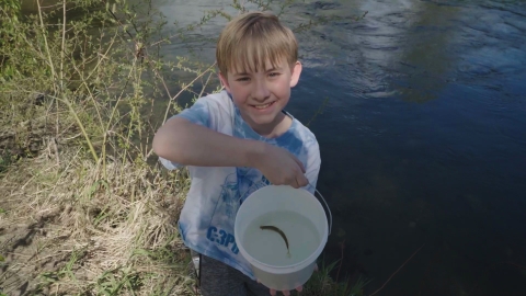 boy in a blue tie-dye shirt stands on the river bank smiling and holding a bucket with a salmon