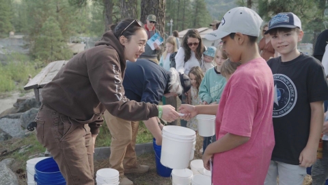 A USFWS staff member hands a small white bucket to a boy in a pink shirt, with a line of happy people in the background 