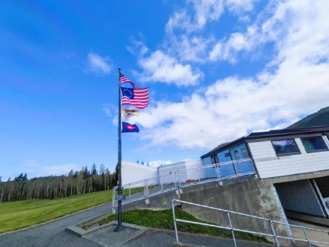 2 US flags, Makah Indian Nation one and fish banner flying on a pole near a building with a ramp, grassy field and trees in the background