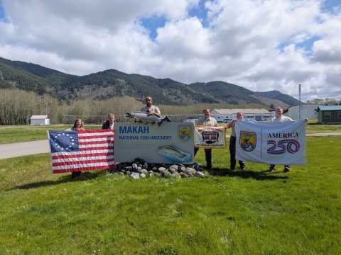 6 people holding Betsy Ross flag, one of the Makah Indian Nation, and USFWS-America 250 one around Makah National Fish Hatchery sign with mountains in background