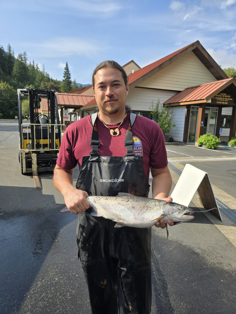 Addison Emerson holding an adult coho salmon at Quilcene National Fish Hatchery. Forklift, facility, and skyline in the background.