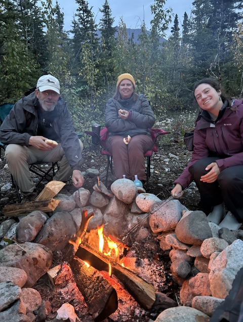 Three people sit around a campfire roasting food.