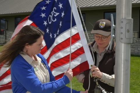 Two people securing a Freedom 250 U.S. flag to a flagpole outdoors