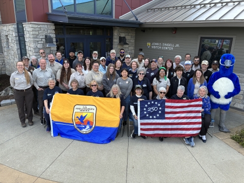 Large group posing outside a building with sign on it saying “JOHN D. DINGELL JR. VISITOR CENTER.” They’re holding USFWS flag and Freedom 250 U.S. flag; blue mascot stands at right