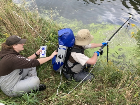 Two USFWS biologists use an eDNA backpack sampler to collect water samplers from the shore.