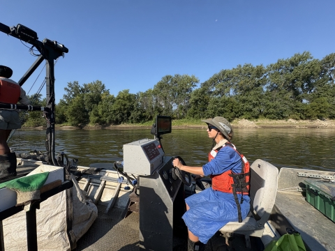 A woman wearing a hat and life vest drives a boat along a river