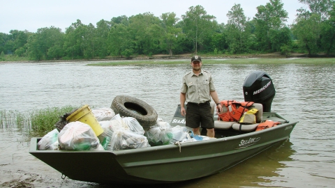 A fish and wildlife service employee stands in a small motorboat filled with trashbags and an old tire