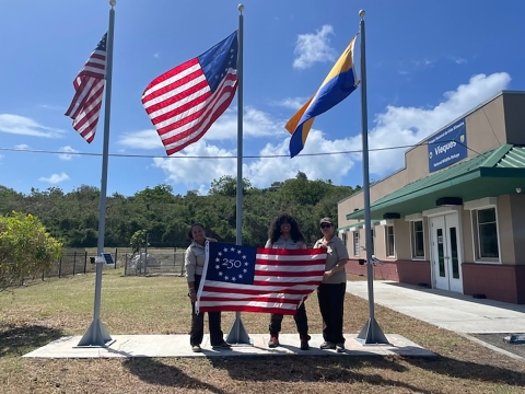Three people hold a Freedom 250 flag in front of a building, with U.S. and FWS flags flying on poles