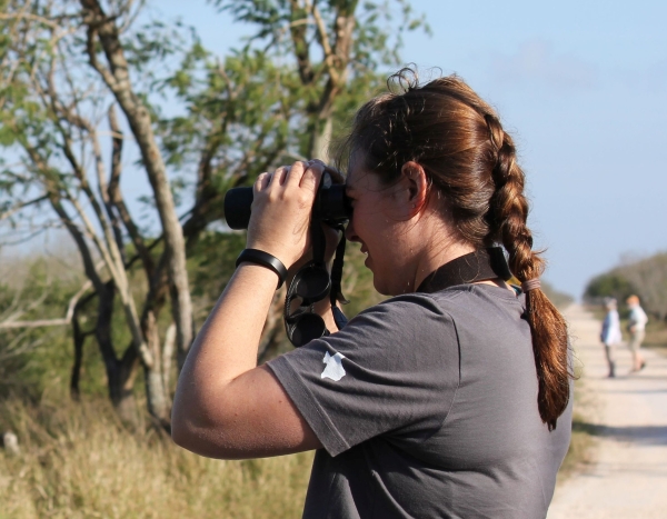 A woman with a braid in her hair looking through binoculars at a scrubby landscape