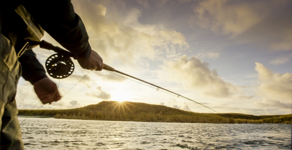 A man standing in a river casting a fly reel with the sun setting behind a distant mountain