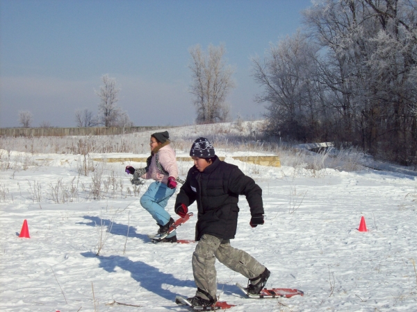Two young adults racing across the snow in snow shoes, which resemble tennis rackets