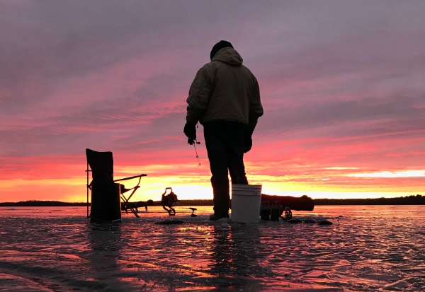 A man in warm-weather gear standing over a hold in the ice with a fishing rod with a colorful sunset in the background