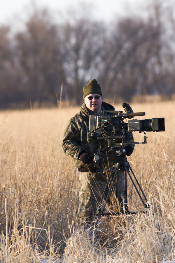 A photographer standing in a open field of grass wearing camo outdoor gear behind a larger camera on a tripod. 