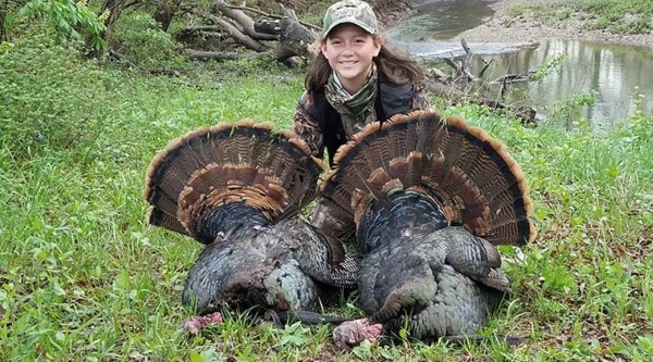 A girl in camouflage crouches on the ground with two large turkeys 