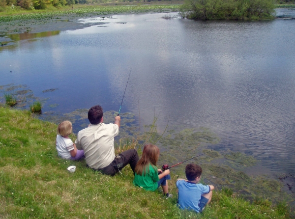 An adult and three children sit on the side of a pond and fish.