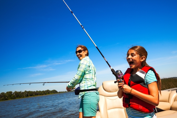 A young girl and her mother laugh as they enjoy fishing on a boat in the middle of a lake in the summer.