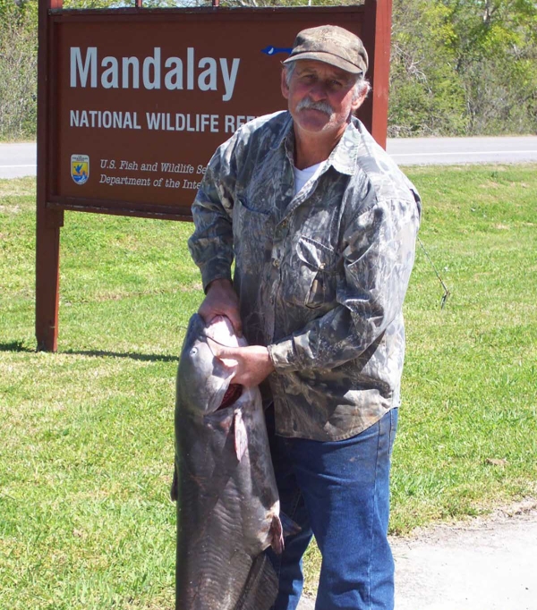 Man holding large fish in front of sign which reads Mandalay NWR