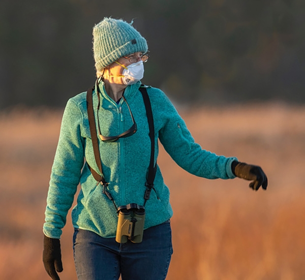 A birder in a mask with binoculars around their neck walks toward the photographer