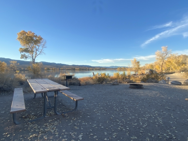 View from campsite number 11 at Pahranagat National Wildlife Refuge. Picnic table, fire ring, grill pit, and a view of Upper Lake with campers in the background.