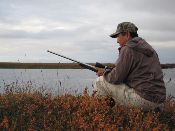 a man crouches on the tundra holding a rifle with a scope