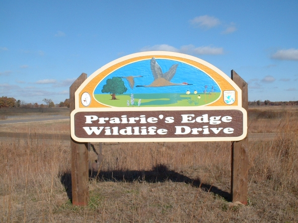 Image of the Prairie's Edge Wildlife Drive wooden sign. This colorful wooden sign has a scene of two sandhill cranes flying in a blue sky over a green landscape, with a tree on the left, wetland in the middle and wildflowers on the right.