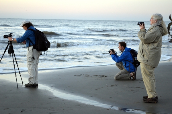 Three people stand at the surf taking pictures of the sunrise on Bulls Island beach.