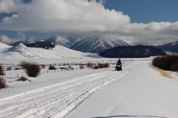 A snowmobiler rides a snowmobile on a snowy road with mountains in the background.