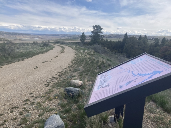interpretive sign in foreground beside gravel road with trees and clouds in the background