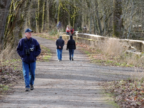 A volunteer in blue uniform and a number of different visitors walk a gravel trail and a boardwalk, wearing jackets and among leafless trees.