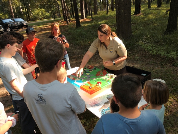 Visitor Services Specialist Amanda Wilkinson uses a 3D model to illustrate how watersheds work to kids. Kids are huddled around a table with the model on it, watching Amanda give the demonstration.