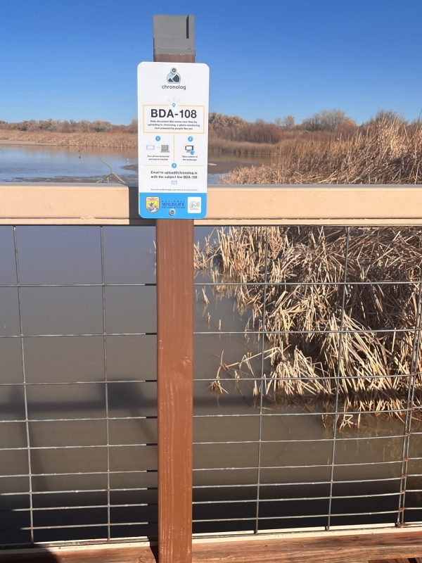 A white and blue sign on a post next to a railing in front of a wetland marsh area with a frame for a mobile phone on top. 