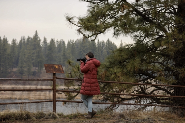 A woman in a red jacket looks through a camera at an overlook