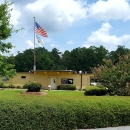 A tan brick building sitting in a grassy courtyard surrounded by trees with a flag pole displaying the American flag and Department of Interior flag.