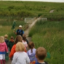 An adult leads a group of young children and moms to a bridge on the wetland in the prairie