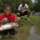Young boy squatting on waters edge holding fish, man in background with fishing rod 