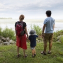 Two adults hold the hands of a child between them and look out at the river from the banks of the Detroit River International Wildlife Refuge.