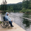 A man in a wheelchair and wearing a hoodie fishing from a platform overhanging a river.